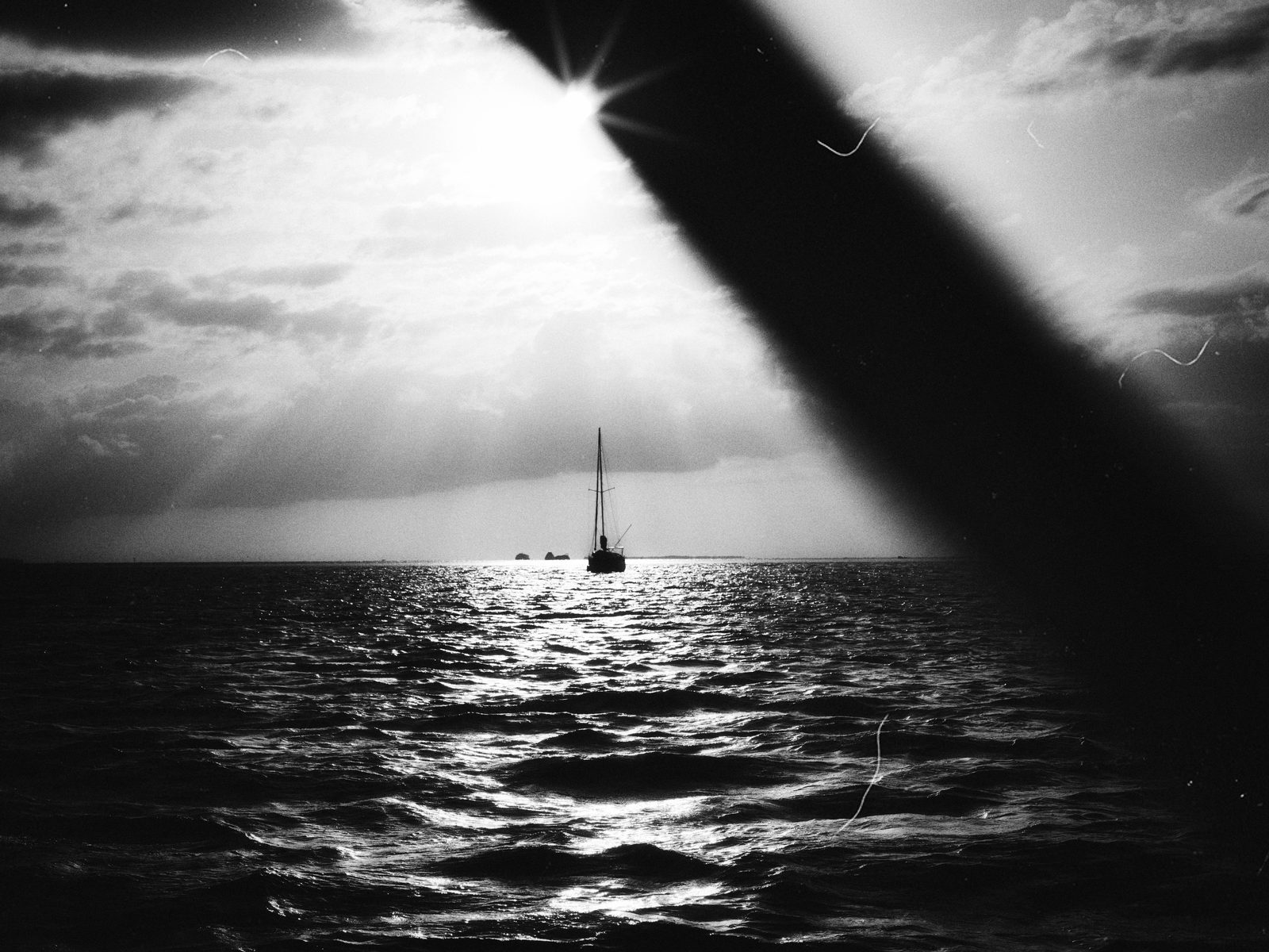 A sailboat at anchor on open water, the sun breaking through cloud