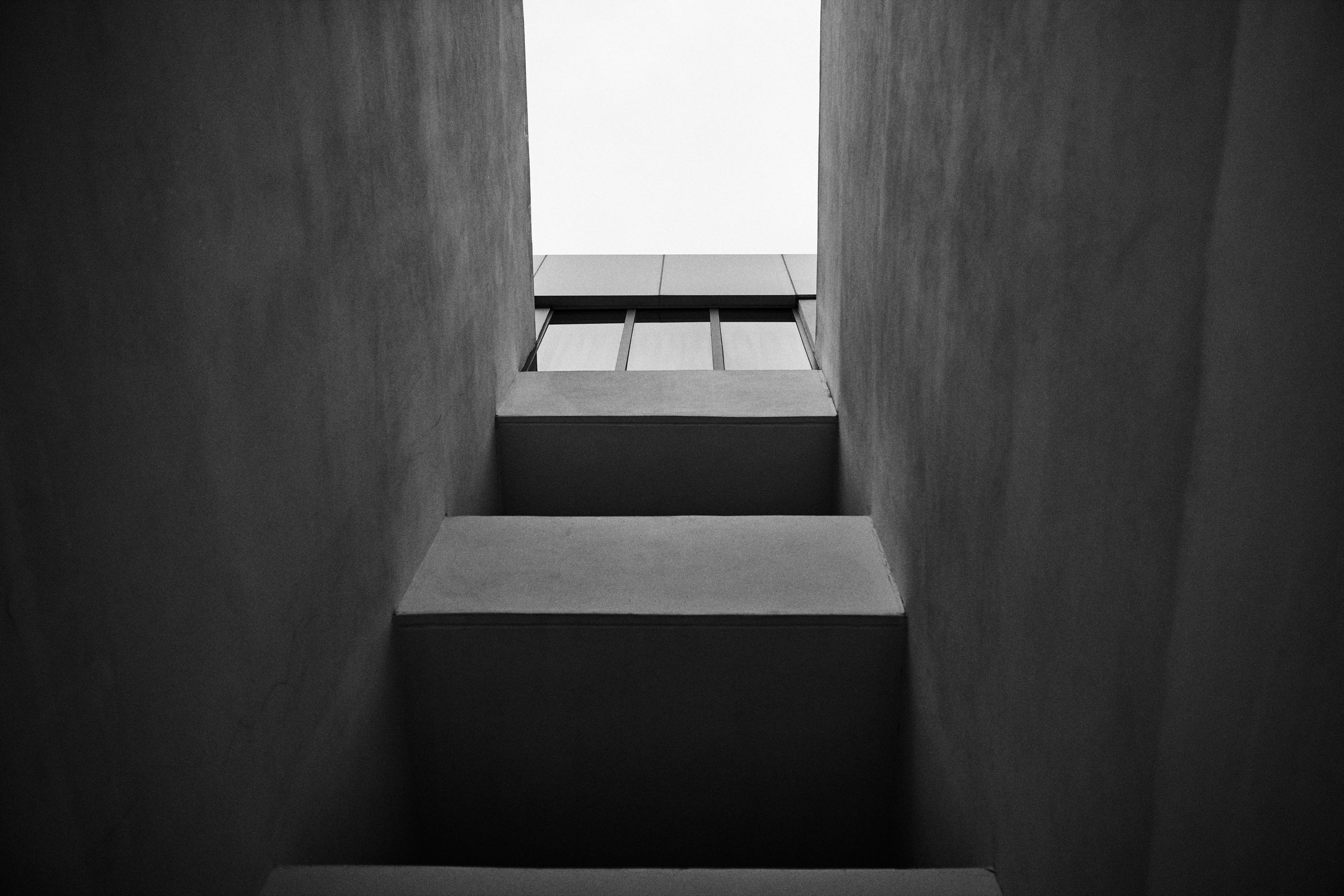 A narrow stair rising between two walls toward a band of windows and open sky