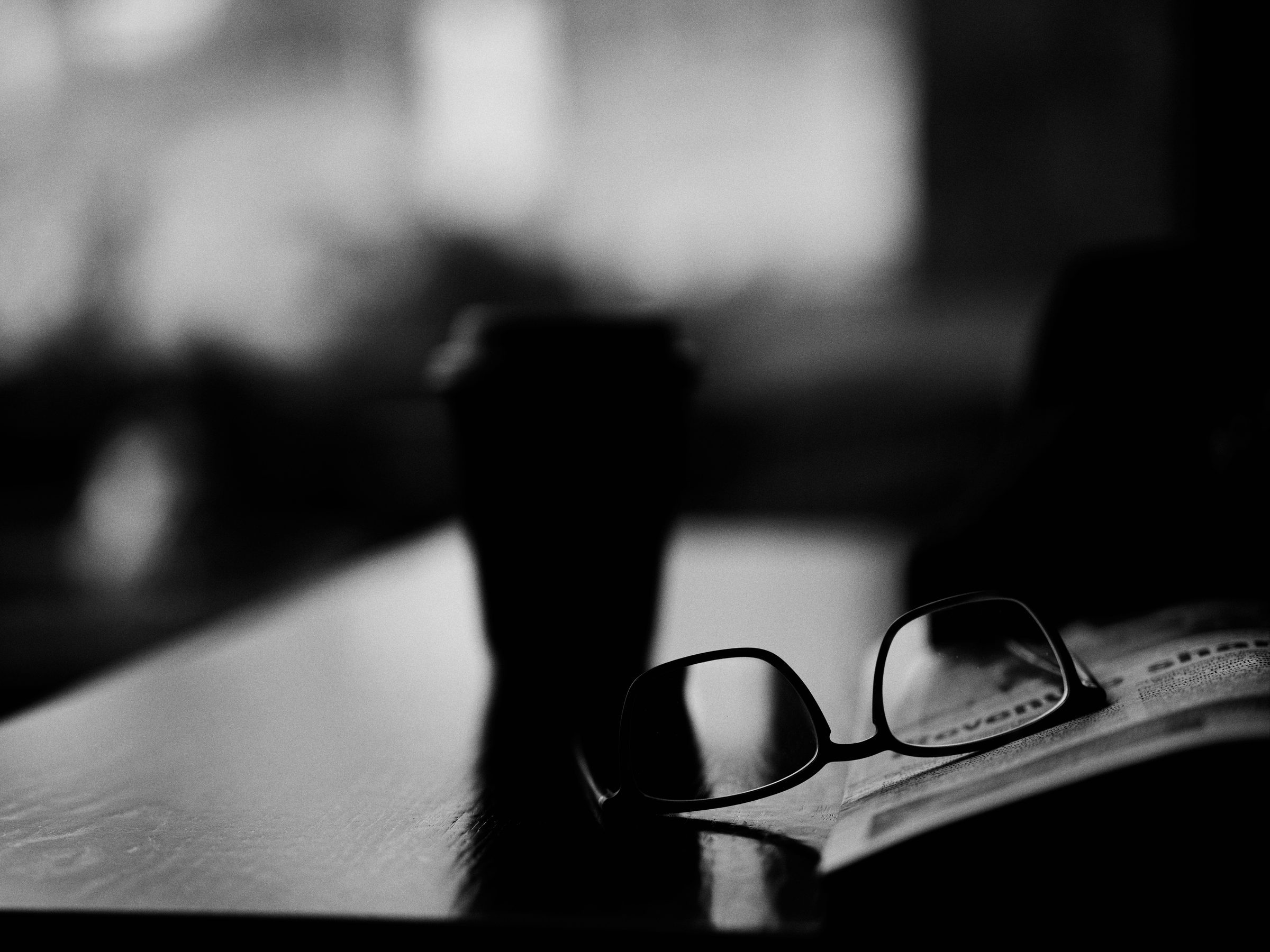 Reading glasses resting on a newspaper beside a cup, window light behind